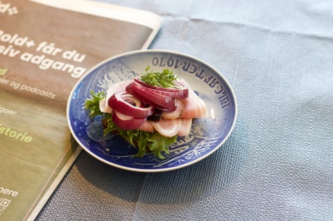 Blue decorated plate with sliced cured meats, fresh greens, and herbs next to a menu on a light fabric surface