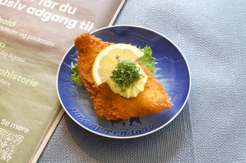 Fried fish fillet on blue plate garnished with lemon slice and green herbs, served on light blue napkin next to menu