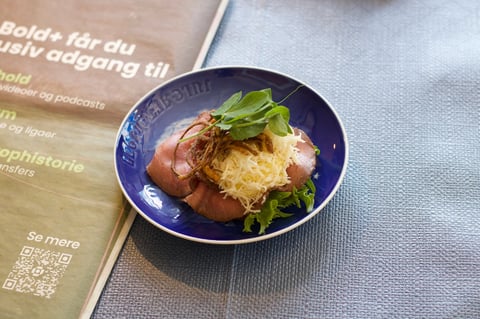 A blue bowl with sliced ham, creamy potato salad, and fresh green herbs sits on a gray surface next to a tan printed menu with text in Scandinavian language