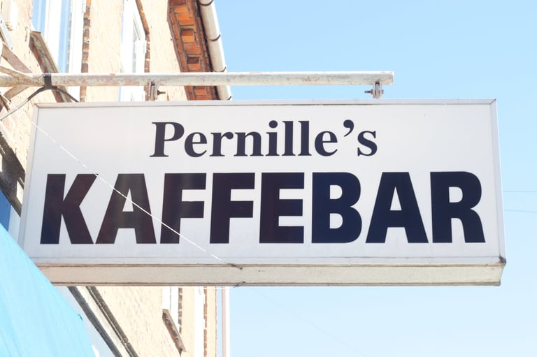 Pernilles Kaffebar hanging storefront sign against a blue sky